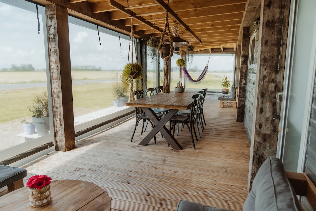 A spacious outdoor veranda features a large wooden dining table surrounded by eight chairs. Hanging plants and a hammock add a touch of greenery, while large glass panels provide unobstructed views of the surrounding fields.