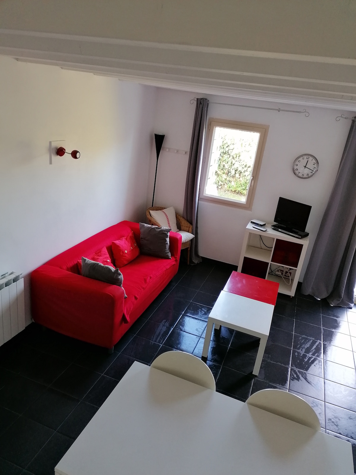 A bright living area features a red sofa adorned with cushions, complemented by a white coffee table. Natural light enters through a window, illuminating the space. A small television is positioned on a white unit, and light grey curtains frame the window.