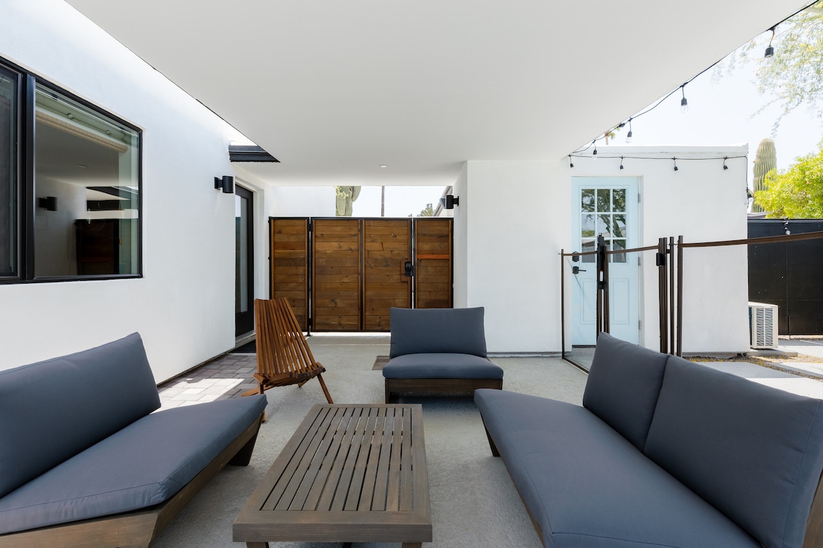 An outdoor lounge area is presented with comfortable gray seating and a wooden coffee table, shaded by a modern overhang. The space features a clean, minimalist design with two wooden gates visible in the background, enhancing the serene atmosphere of the patio.