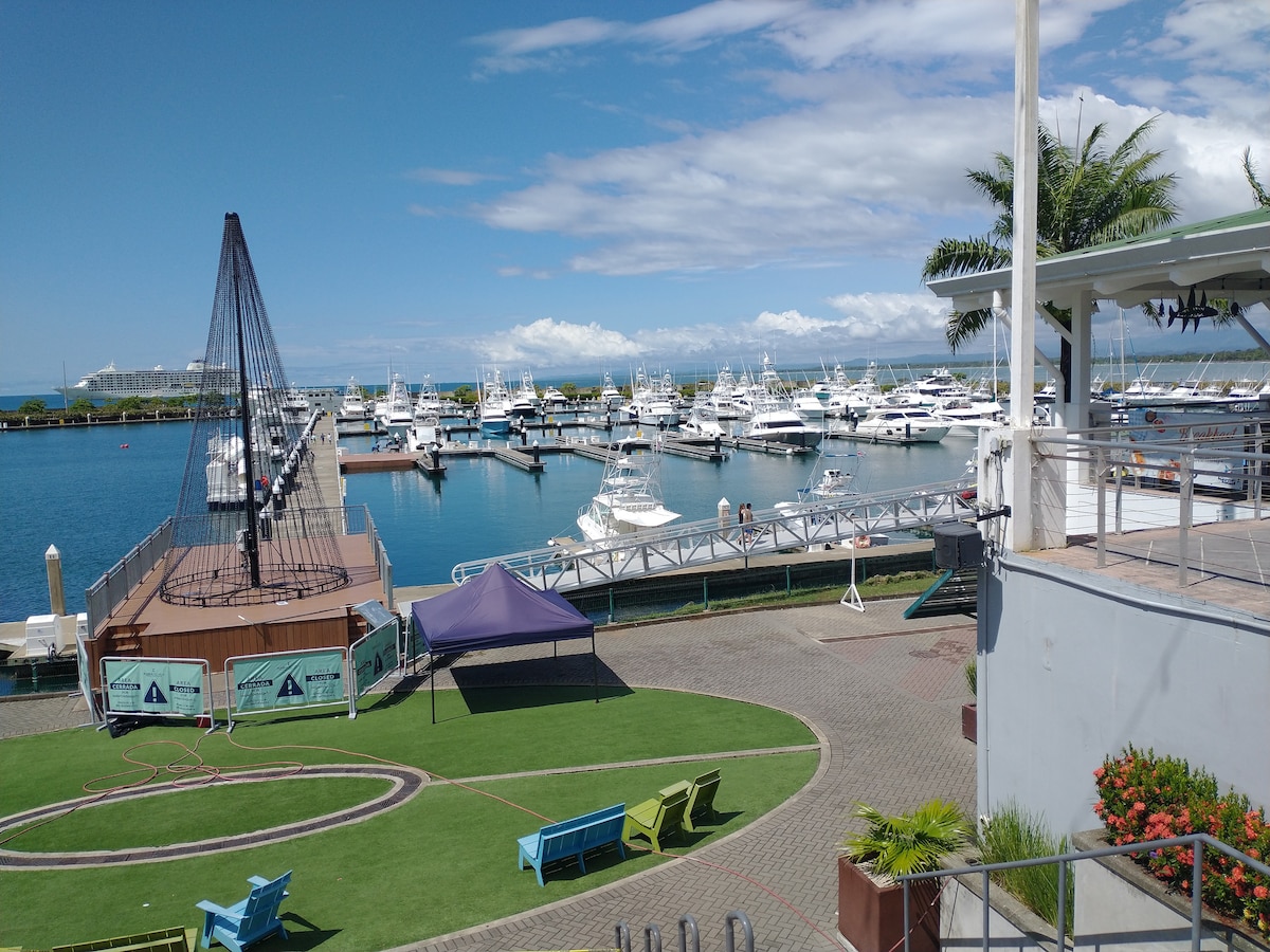 A marina scene showcases a collection of moored yachts and boats along a calm, azure waterway. Lush green areas surround the dock, complementing the bright blue sky. A modern structure is visible in the foreground, enhancing the harbor's vibrant atmosphere.