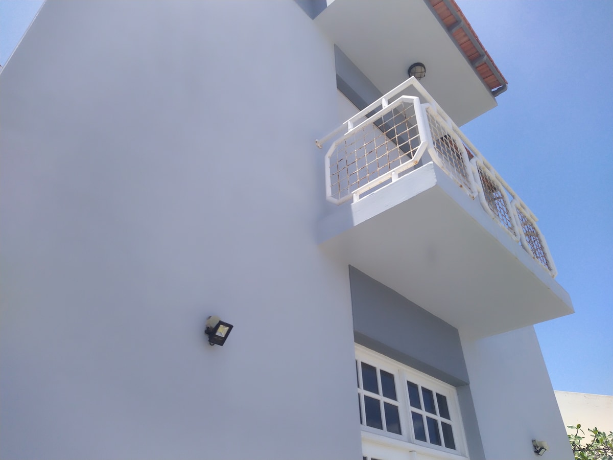 A white exterior portion of a two-story building is shown, with a balcony extending from the second floor. A large window with multiple panes is visible, and modern lighting fixtures are mounted on the walls, providing additional illumination.