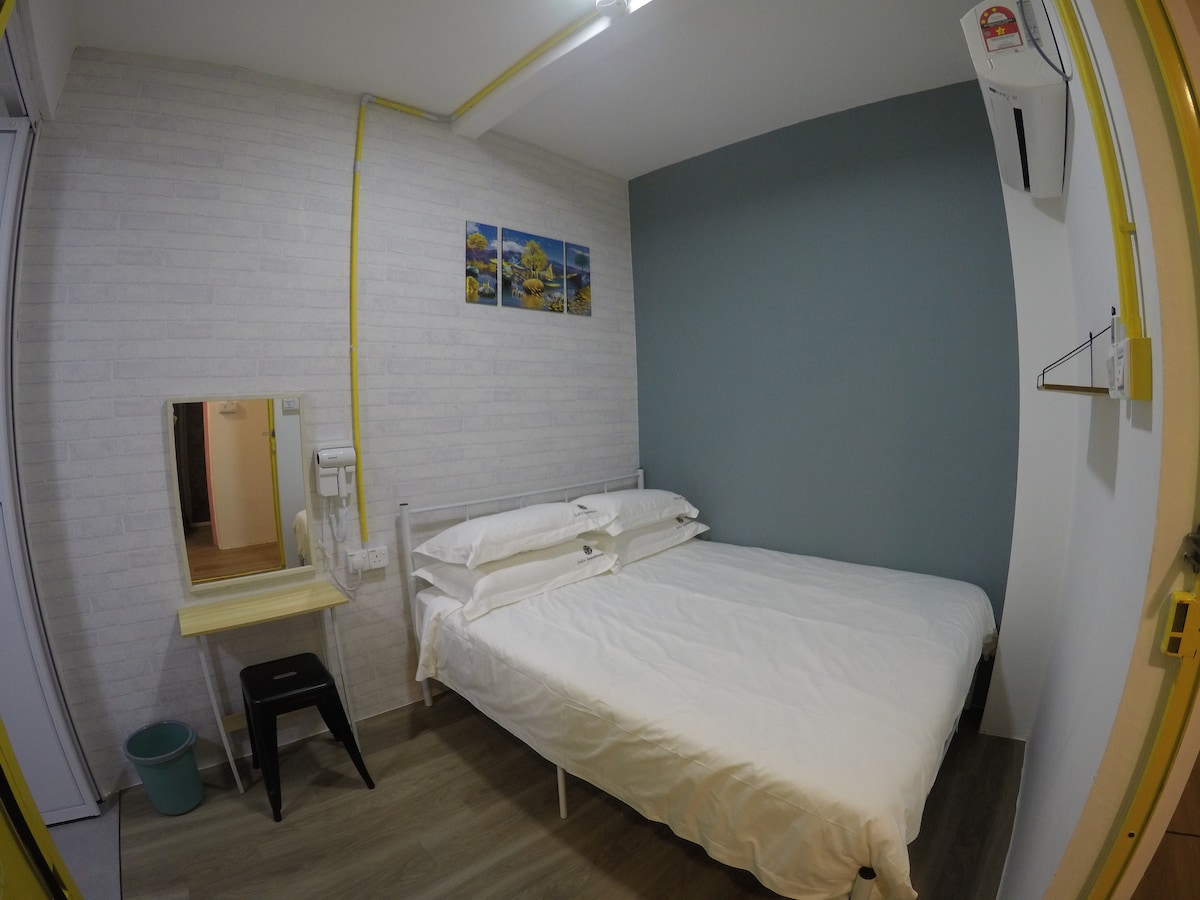 A well-lit bedroom features a spacious bed with crisp white linens. A small work desk and a mirror are positioned nearby. Light-colored walls are complemented by a blue accent wall, creating a subdued environment. A wardrobe and a small wastebasket are also visible in the room.