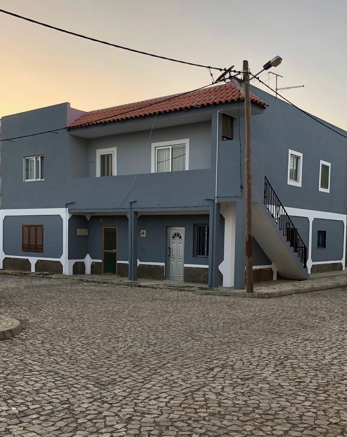 A two-story building is depicted, featuring a gray exterior with white accents. The entrance is clearly marked, and a staircase is visible on the side. Cobblestone paving surrounds the property, creating a quaint streetscape.