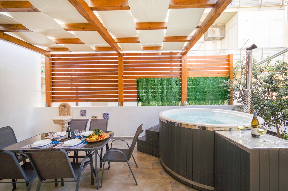An outdoor space is presented featuring a hot tub and a dining table set for four. The area is shaded by wooden beams and features decorative wall elements, offering a cozy atmosphere for relaxation and dining.