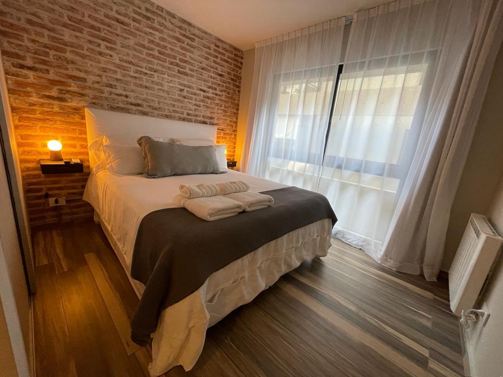 A serene bedroom features a spacious bed with neutral bedding and a cozy throw. Soft lighting is provided by bedside lamps, and natural light fills the room through large windows draped with sheer curtains. Exposed brick walls add a unique texture.
