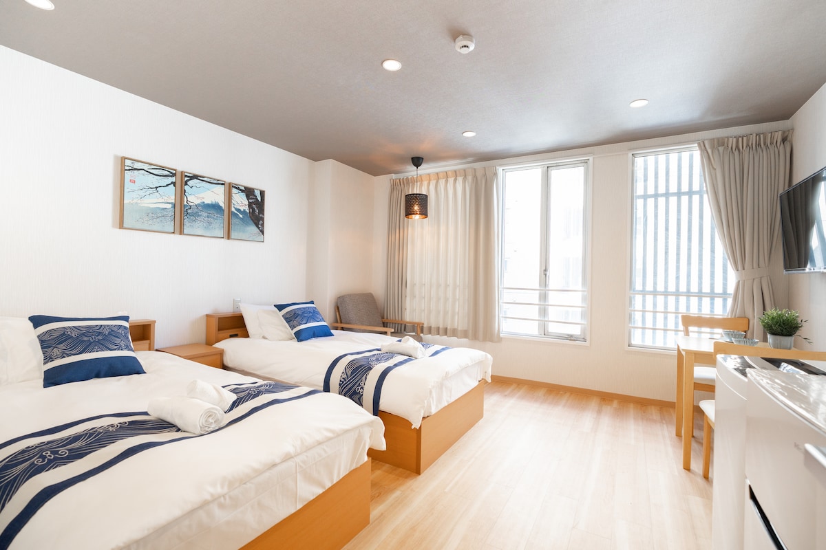 A spacious guest room features two single beds with blue accent pillows, paired with a small seating area and a wooden chair. Natural light enters through two large windows, which are covered with sheer curtains. Minimalistic artwork adorns the wall.