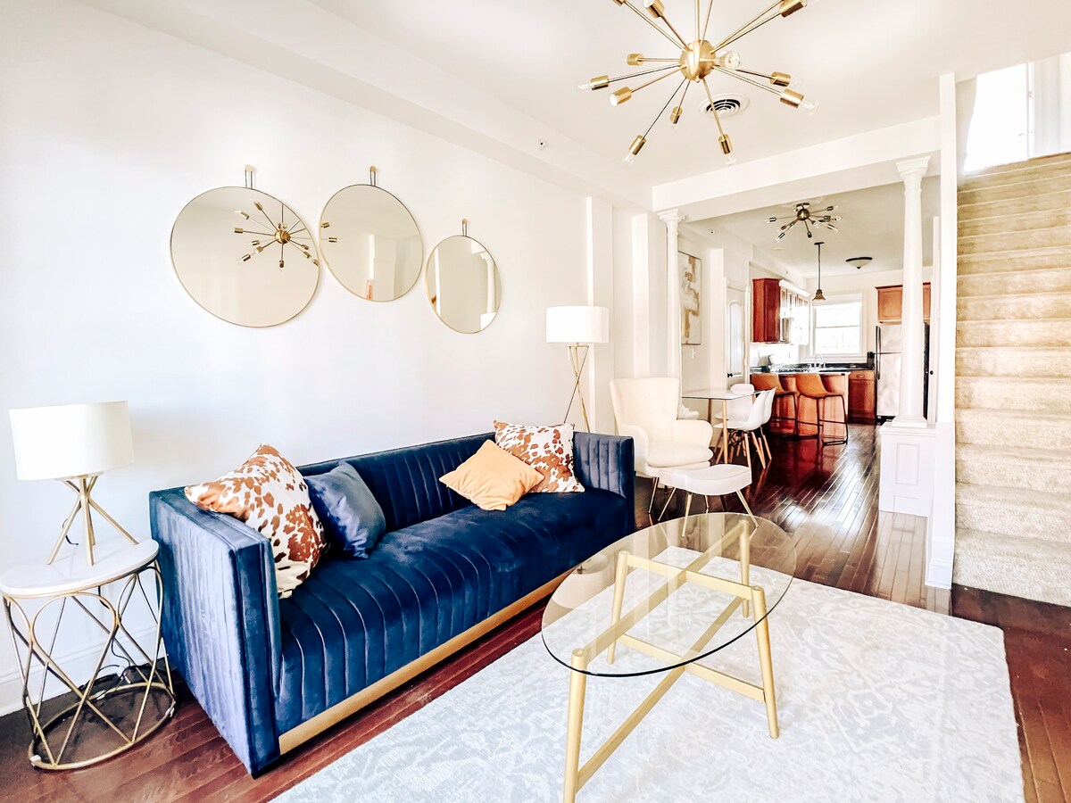 A bright living area features a navy blue sofa with colorful cushions, accompanied by two stylish side tables and a glass coffee table. Three round mirrors are mounted on the wall, reflecting light into the space. Staircase access is visible on the right side.