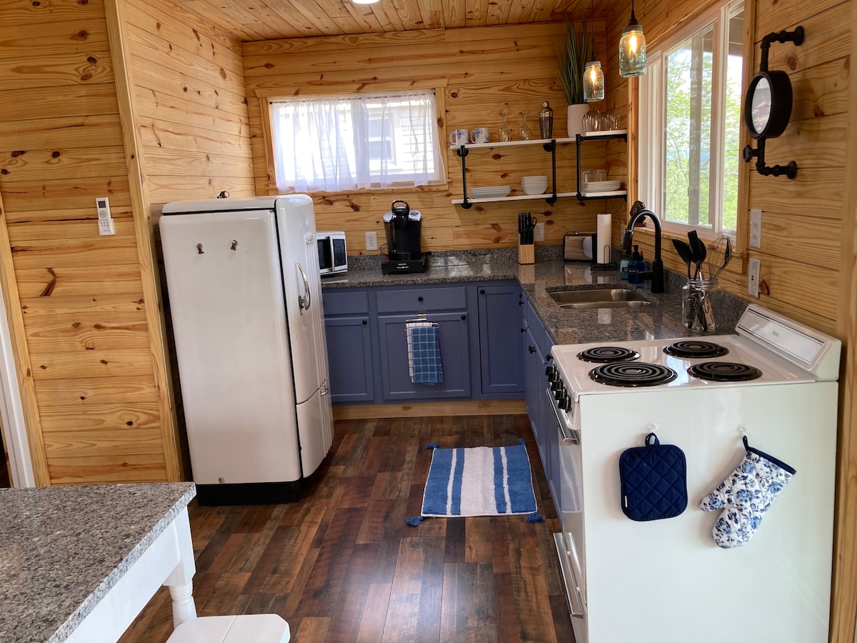 The kitchen features wooden paneling, granite countertops, and vintage appliances, including a 1950's refrigerator. Natural light floods the space through large windows. Cooking equipment and utensils are neatly arranged, with a coffee machine and a striped rug adding functional touches.