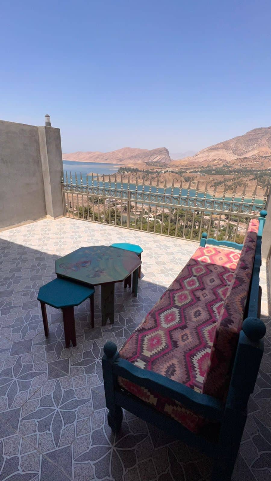 A balcony area features a vibrant patterned sofa and a uniquely shaped table, set against a stunning view of the lake and mountains. The tiled flooring adds texture, while the wrought iron railing offers safety and a framed perspective of the surrounding natural beauty.