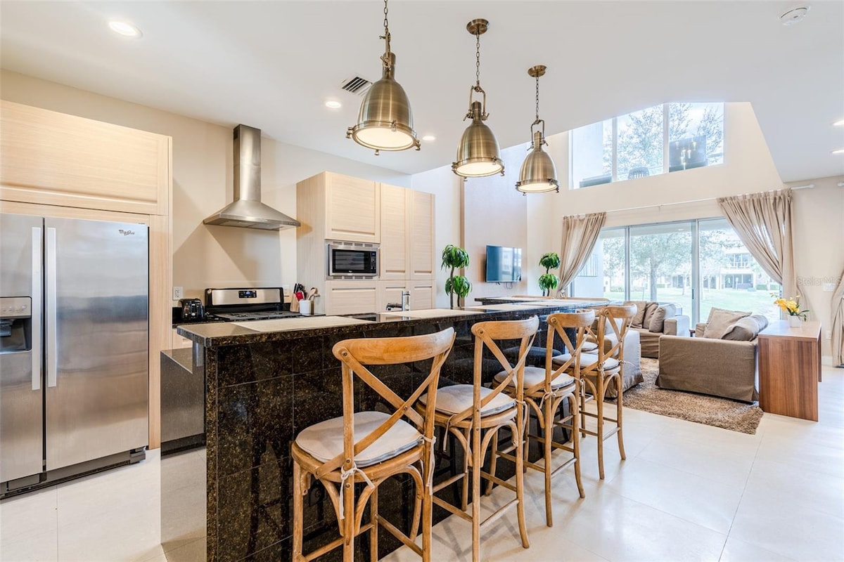 A modern kitchen area features a large dark countertop with tall wooden chairs positioned around it. Stainless steel appliances and a range hood are visible. Natural light fills the space through a high window, illuminating the adjacent living area furnished with a sofa.