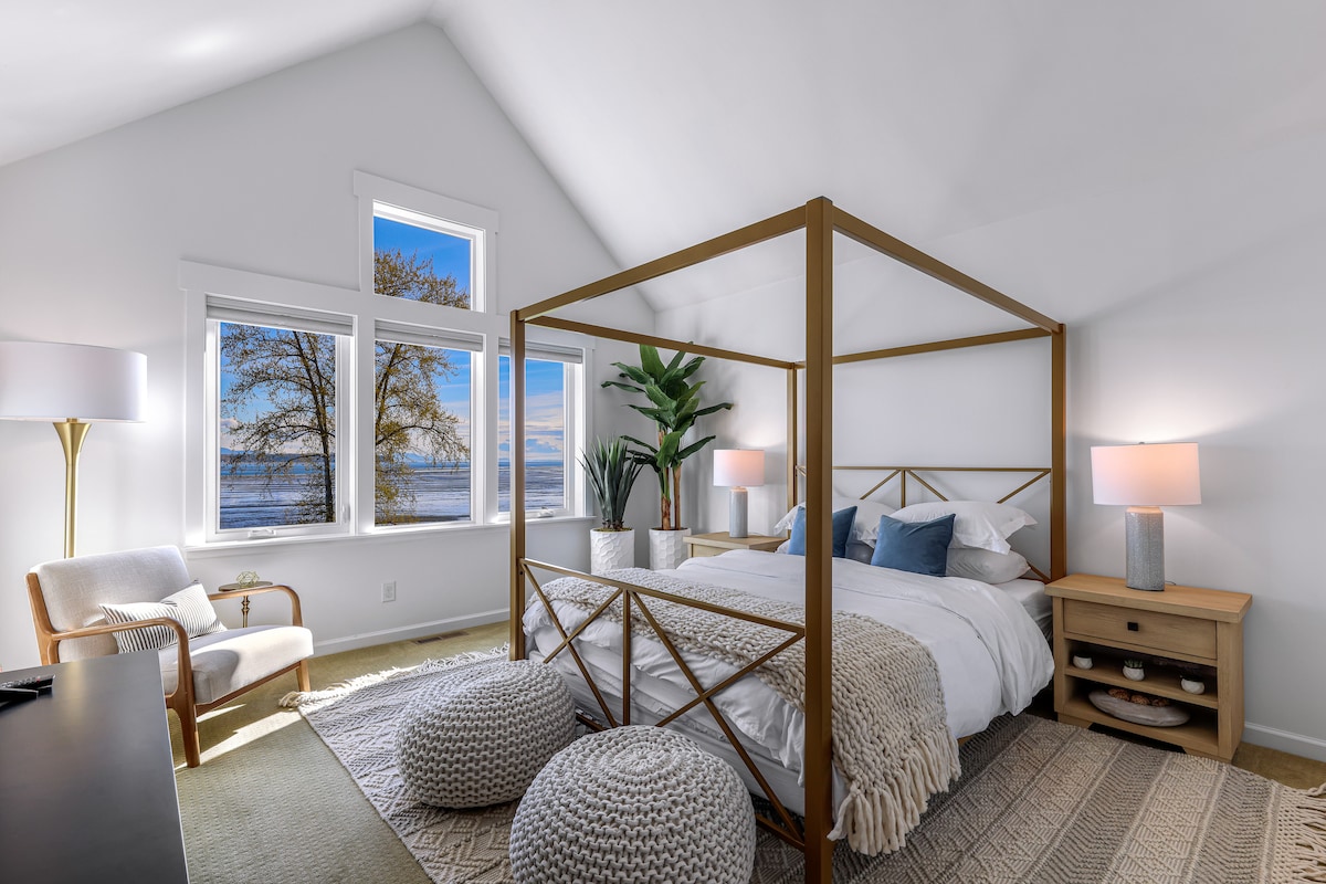 The primary bedroom is bright and airy, featuring a spacious four-poster bed under high ceilings. Large windows provide uninterrupted views of Birch Bay. Soft natural light illuminates the room, complemented by bedside lamps and a cozy seating area with a chair and decorative poufs.