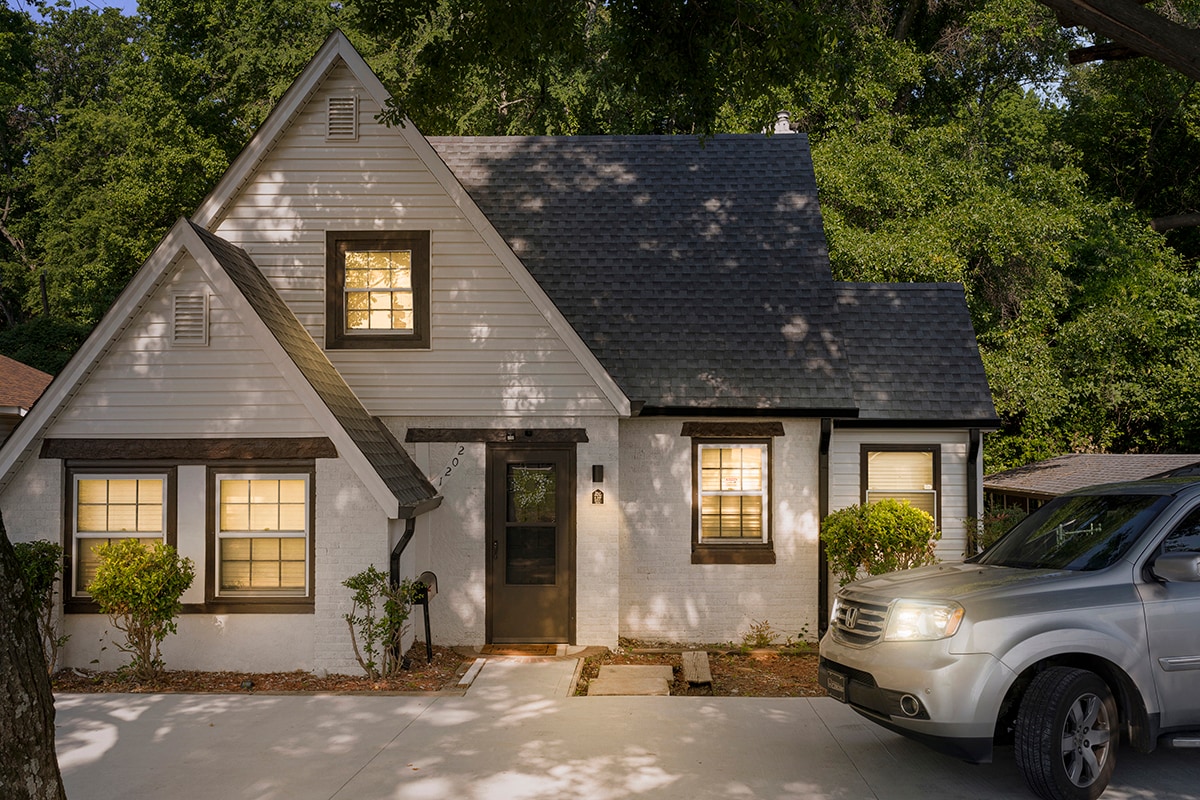 The exterior of the cottage-style home is depicted, showcasing a steep roof and distinct architectural features. The entrance is positioned on the right side, flanked by neatly trimmed shrubs. A spacious driveway accommodates vehicle parking. Soft evening light filters through the windows.