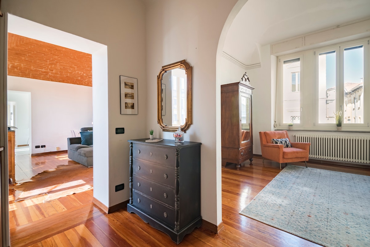 An inviting entryway features a dark wooden dresser and a vintage mirror. An arched passage leads to a light-filled living area, where a comfortable armchair sits beside a window. Hardwood floors extend throughout, reflecting the natural light from the tall windows.