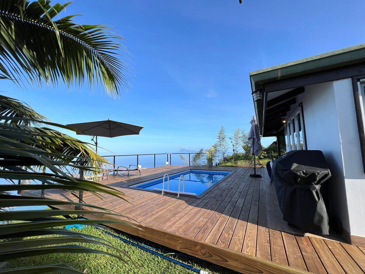 A wooden deck surrounds a heated swimming pool, offering unobstructed views of the sky and distant mountains. A shaded umbrella provides a resting area, while a grill is positioned nearby, enhancing outdoor dining options in a tranquil setting surrounded by greenery.