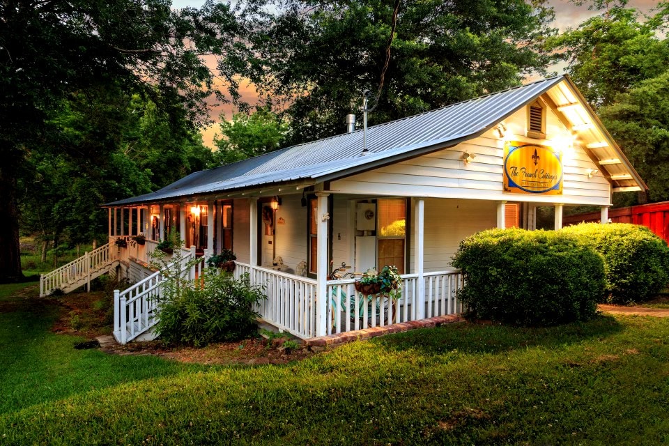 A charming cottage with a wraparound porch is positioned on a lush green lawn. Warm light spills from the windows, revealing a welcoming atmosphere. The exterior features a classic metal roof, while well-kept shrubs and ornamental plants add to the inviting landscape.