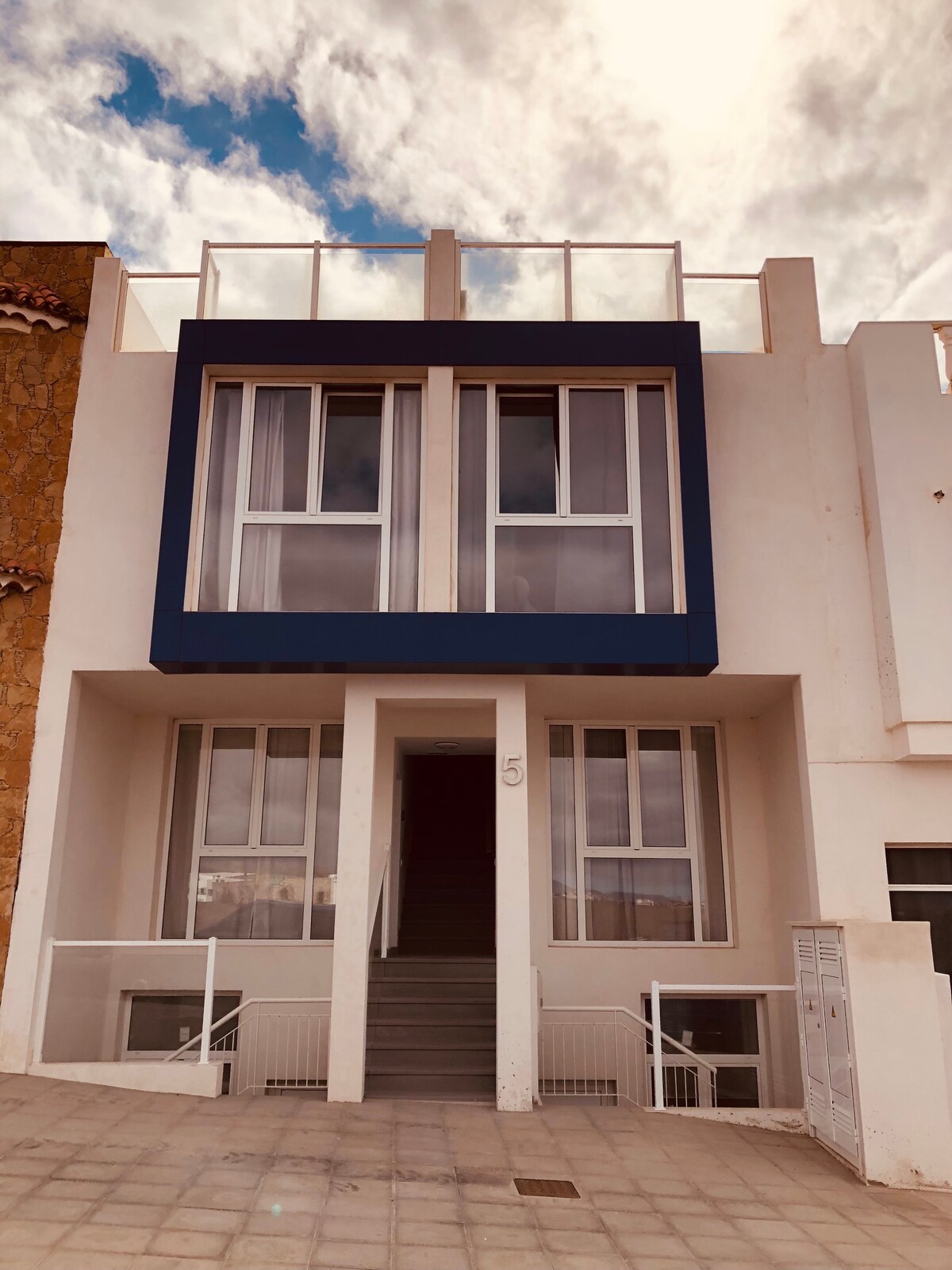 The exterior of a newly constructed building is showcased, featuring a modern façade with large windows bordered by a blue frame. A central entrance leads to a staircase. The surrounding area is paved, with a few steps leading up to the entrance.