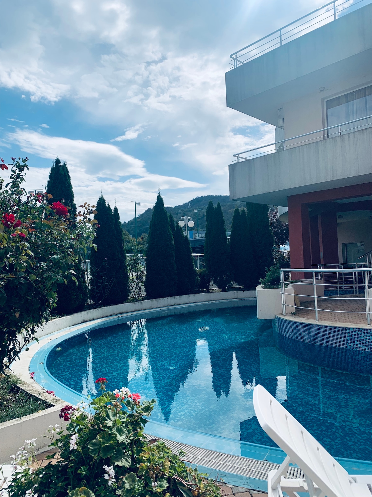 A curved swimming pool is surrounded by well-maintained greenery and flowering plants. Tall coniferous trees create a natural backdrop, reflecting their silhouettes on the water's surface. The building's modern exterior features a mix of warm and neutral tones, contributing to a serene outdoor environment.