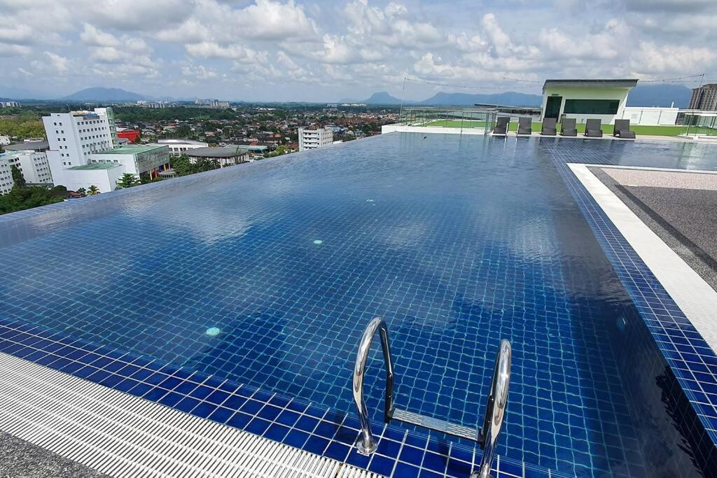 An infinity swimming pool is positioned on the rooftop, showcasing a reflective surface that merges with the skyline. Lounge chairs are situated nearby, inviting relaxation. The backdrop features a panoramic view of the cityscape and distant mountains, under a clear blue sky with scattered clouds.