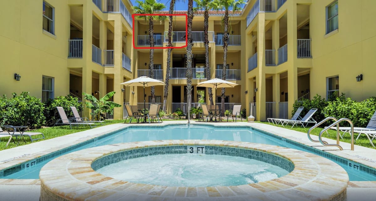 The image showcases a central courtyard featuring a swimming pool and hot tub. Surrounding greenery includes palm trees, and lounge chairs are placed around the pool area. The balcony of the third-floor unit is highlighted, offering a view of the tranquil setting.