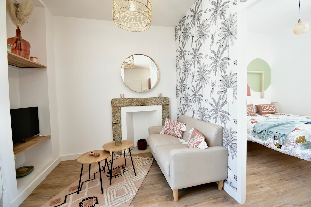 A living area is presented with a light-colored sofa adorned with decorative cushions. Two wooden tables are positioned in front of the sofa, alongside a TV mounted on a wooden shelf. A feature wall with palm leaf patterns contrasts against the bright space.