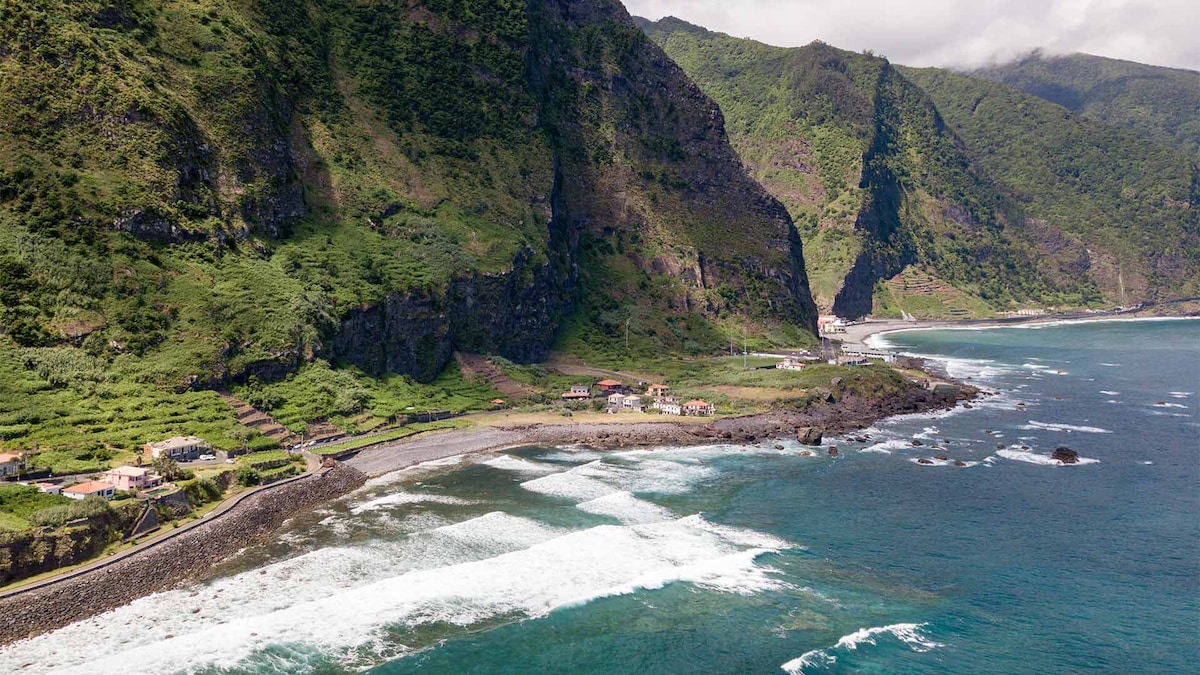 A scenic coastal view showcases rugged cliffs meeting the ocean, with waves gently crashing on the rocky shoreline. A few small buildings are visible along the beach, surrounded by lush greenery and mountains in the background, providing a tranquil natural landscape.