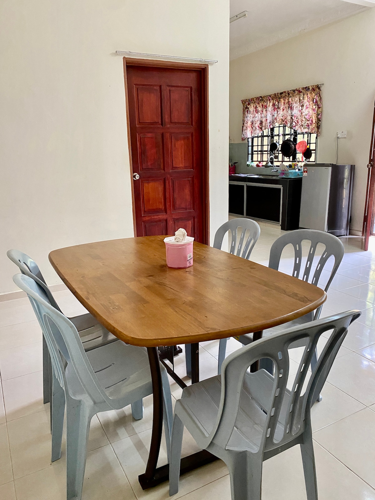 A wooden dining table is surrounded by six light blue plastic chairs. The space is well-lit, with a door visible in the background and a window adorned with floral curtains. A small pink container is placed on the table.