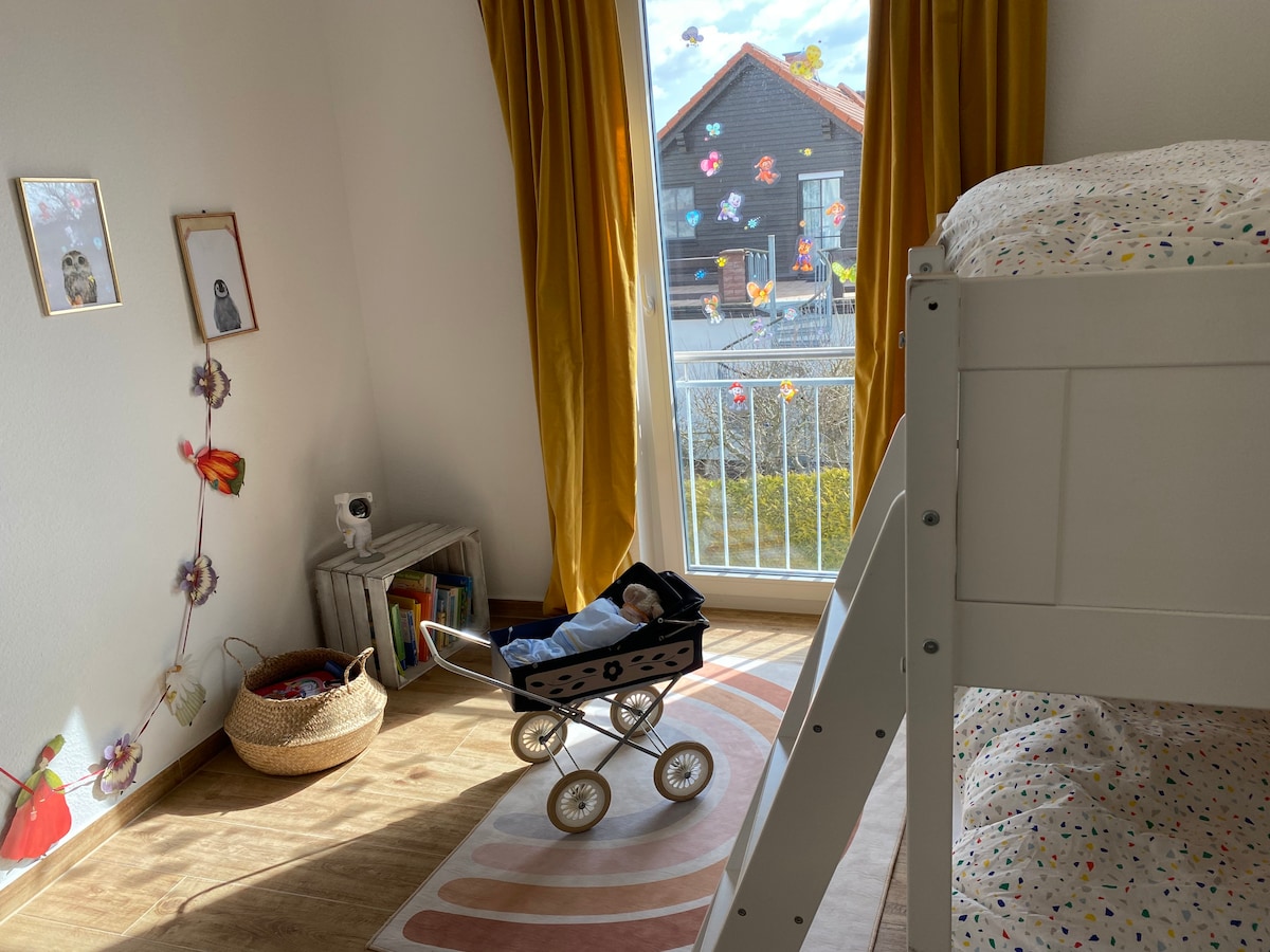 The children's room features a bunk bed, a vintage doll stroller, and a soft rug. Natural light fills the space through a large window, complemented by cheerful curtains. Decorative elements include framed pictures on the wall and a basket for toys, creating a playful atmosphere.