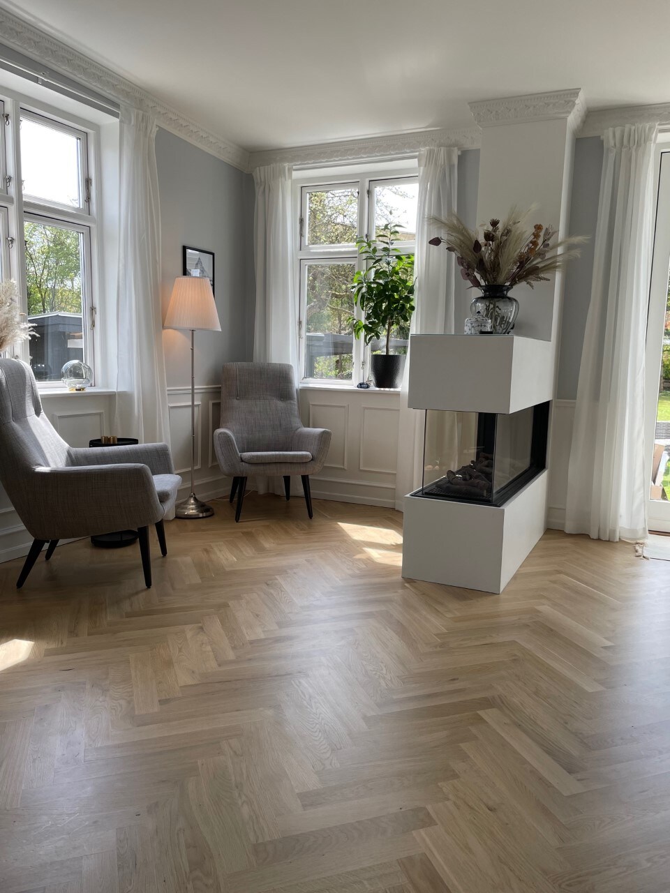 A bright living area features large windows allowing natural light to fill the space. Two upholstered chairs are positioned near a modern, minimalist fireplace. Light wooden flooring complements the soft neutral tones of the walls, while a decorative plant adds a touch of greenery.