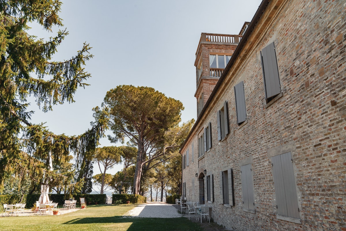 The exterior of the historic country house features a well-preserved rustic stone facade, complemented by grey shutters. The surrounding landscape includes tall trees and outdoor seating areas, offering a serene environment. Sunlight filters through the foliage, creating a peaceful setting for relaxation.