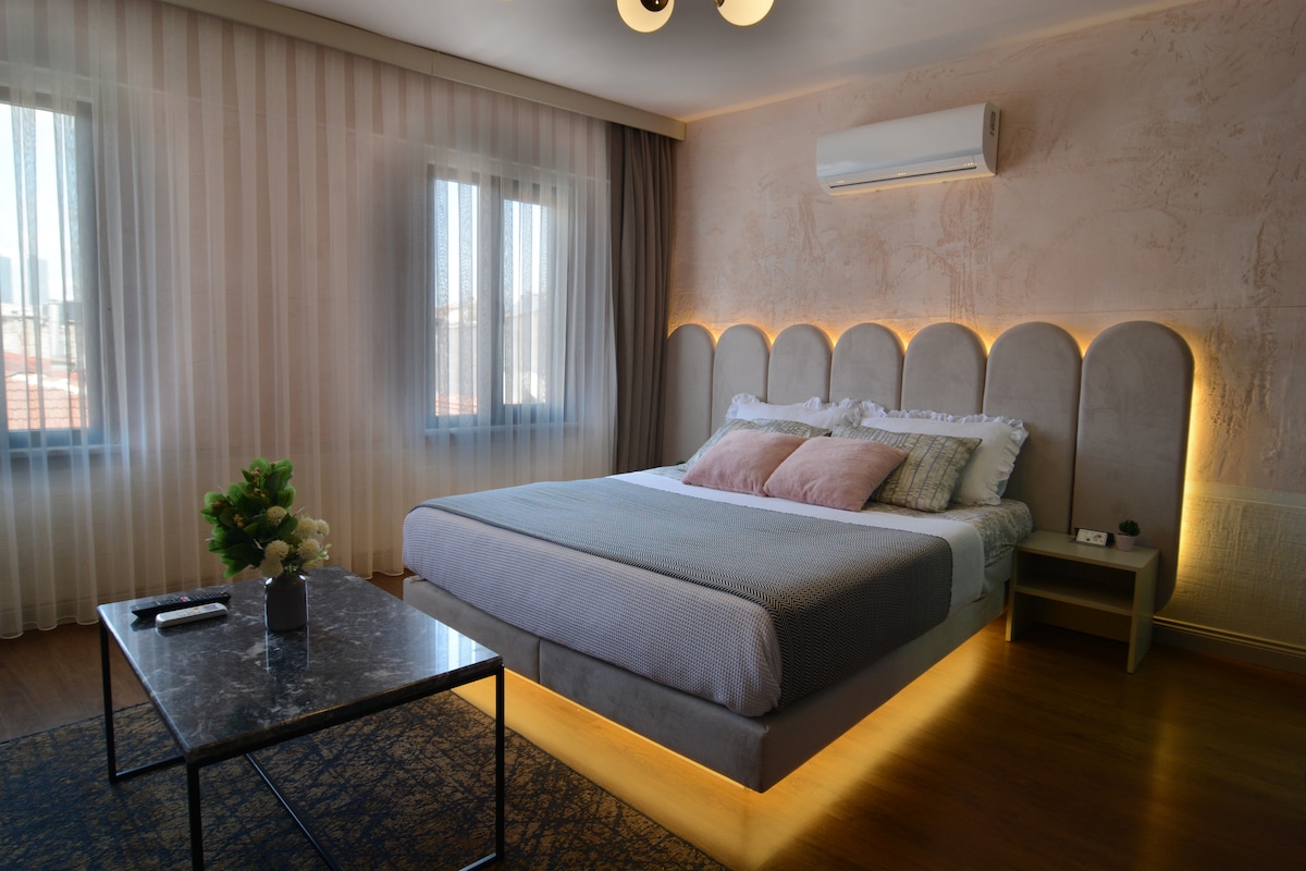 A spacious bedroom is illuminated by soft lighting from the bed's base, which features a unique headboard design. Two windows are dressed with sheer curtains, allowing natural light to enter. A small table with a decorative plant and a remote control rests beside the bed.