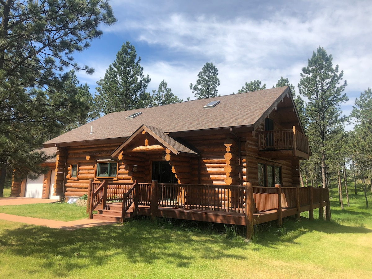A log cabin home is nestled among tall pines, showcasing a wrap-around deck with wooden railings. Large windows invite natural light, and the structure's deep brown logs contrast with the surrounding greenery. A pathway leads to the entrance, highlighting the peaceful outdoor setting.