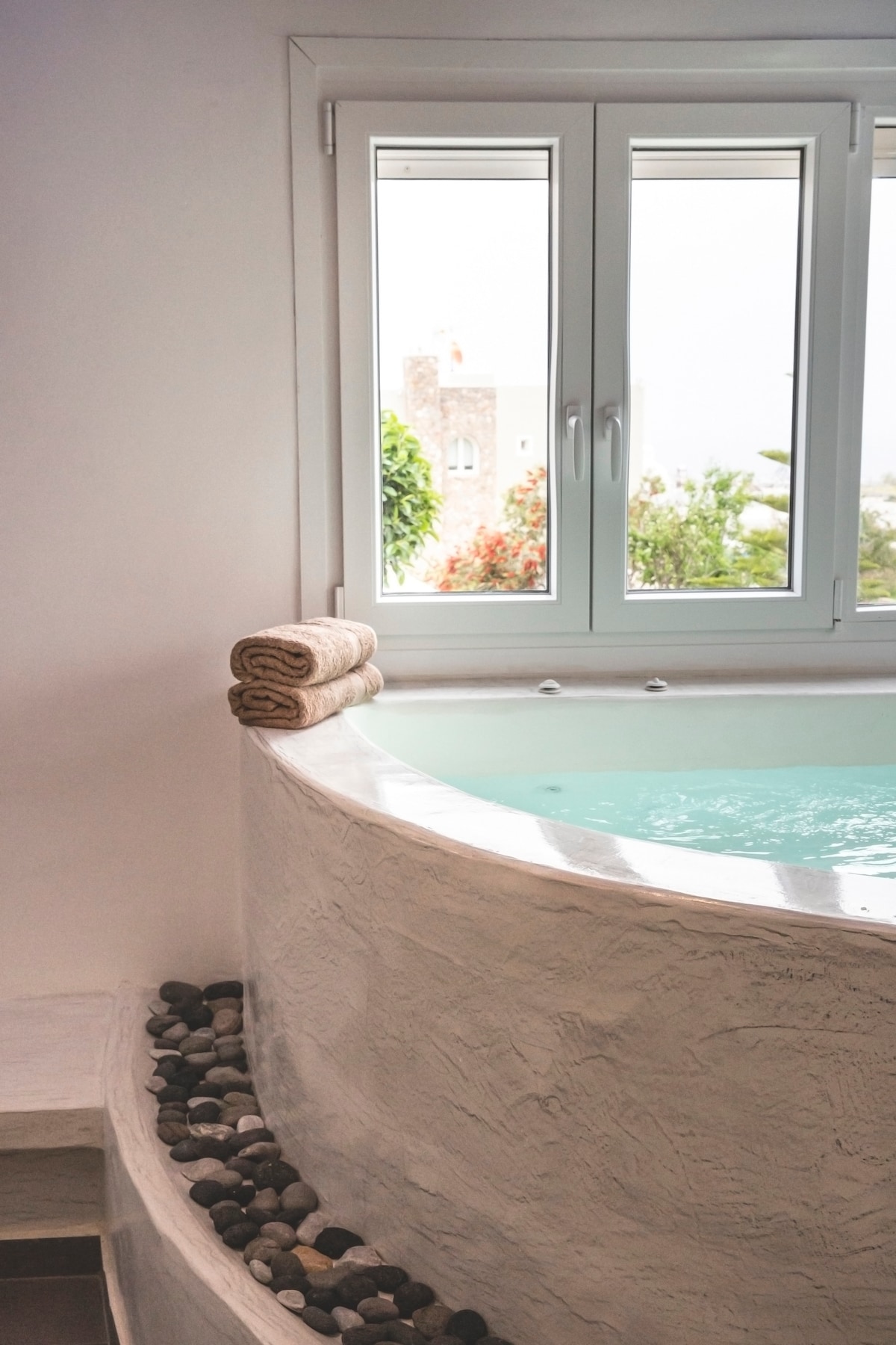 A soothing outdoor hot tub is framed by a white wall and a window, showcasing gentle water. Rolled towels rest on the edge, and smooth pebbles line the base, creating a serene environment. Greenery is visible outside, enhancing the calming atmosphere.