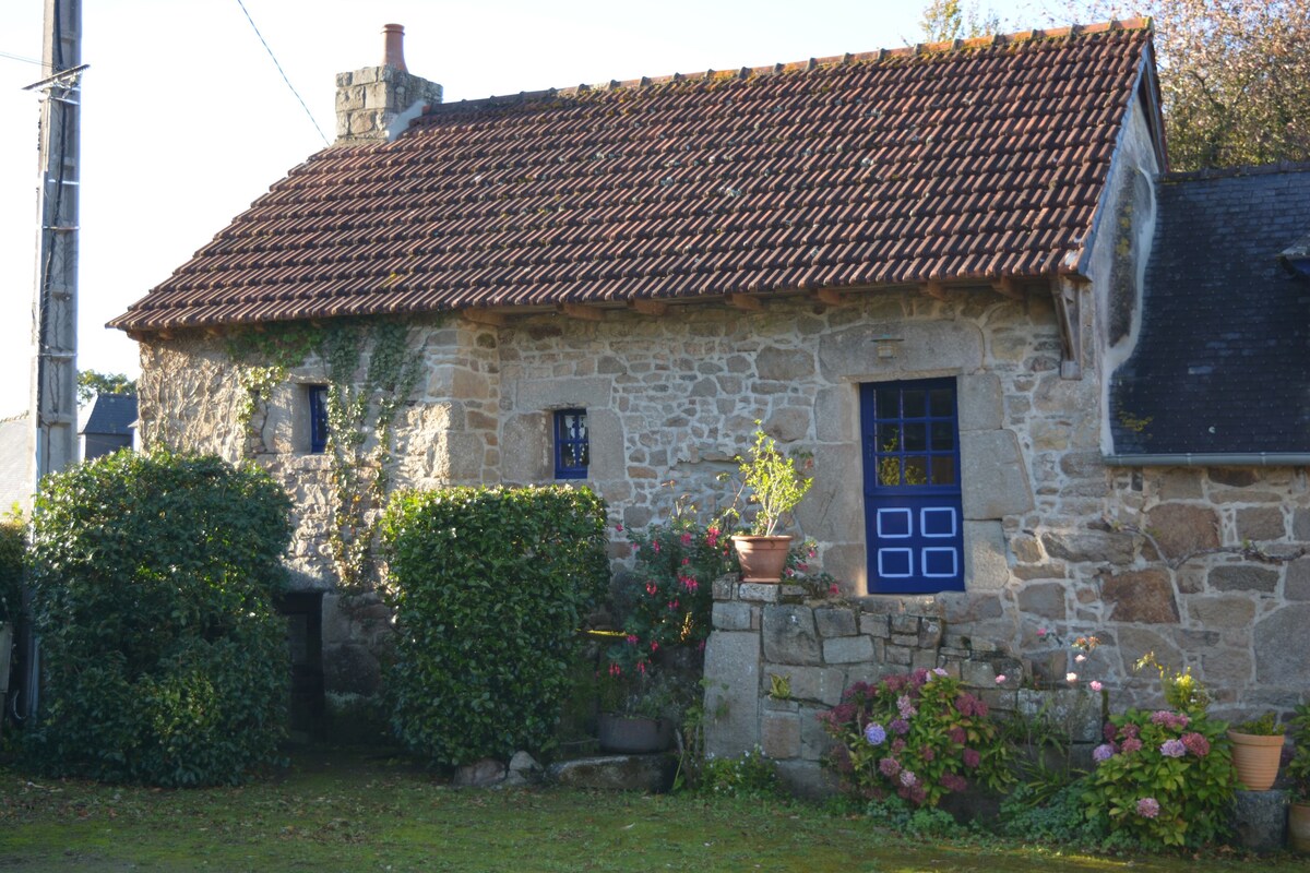 A traditional stone cottage is framed by lush greenery and colorful blooming flowers. The structure features a distinctive blue door and rustic red roof tiles, emphasizing its charming character. Natural light softly illuminates the exterior details, creating a serene and peaceful setting.