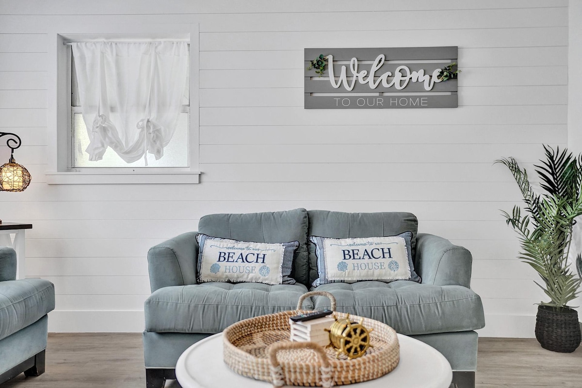 A cozy living area is highlighted by a soft blue sofa adorned with 'BEACH HOUSE' pillows. A decorative light fixture hangs nearby, while a welcoming sign is mounted on the wall. Natural light filters through the sheer curtains at the window, enhancing the space.