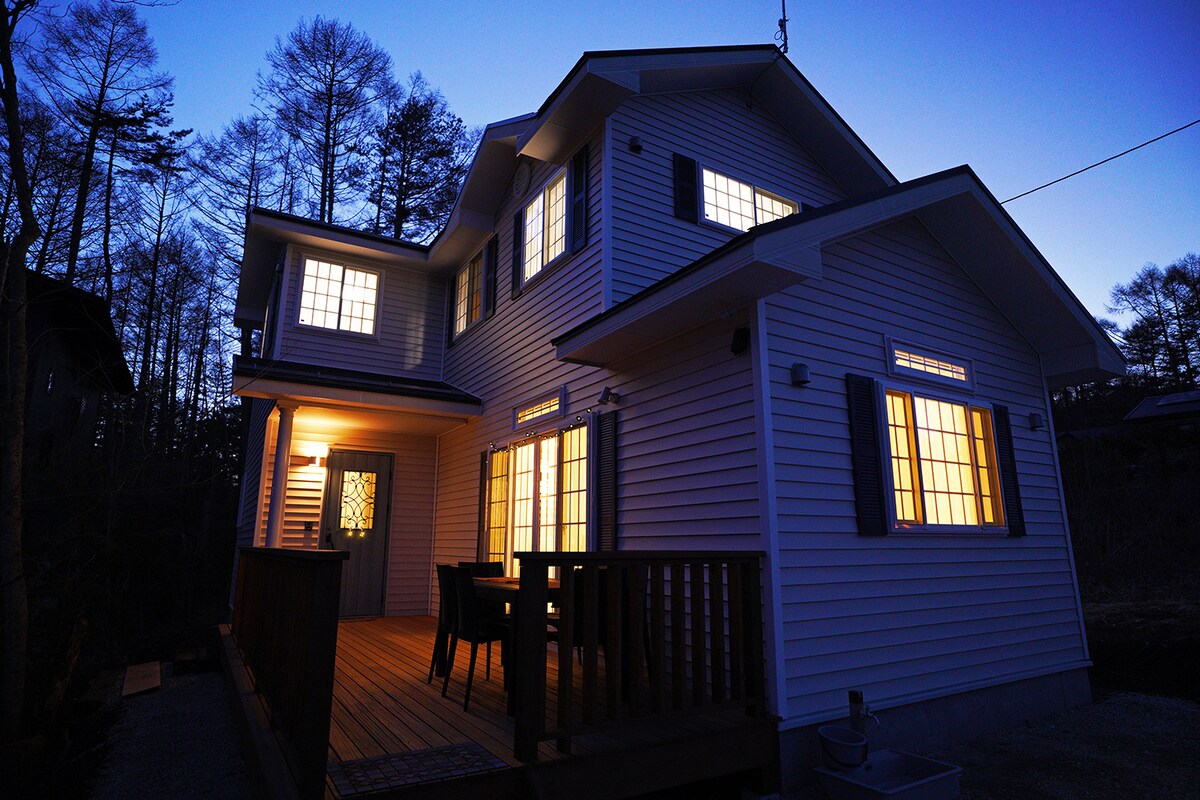 The exterior of the white house is illuminated at dusk, with warm light glowing from the windows. The structure features a wooden deck in the foreground, surrounded by trees that create a natural backdrop.