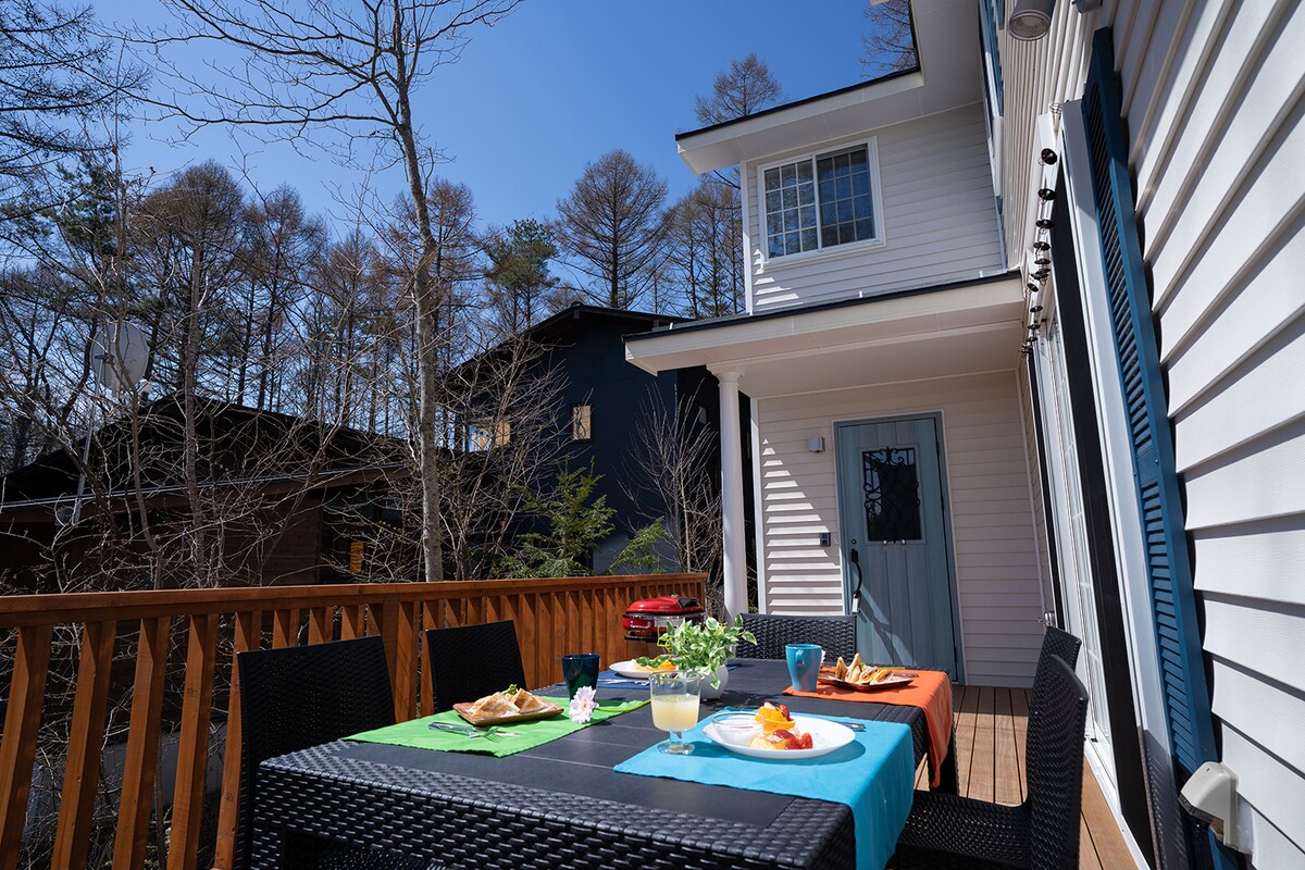 A spacious wooden deck is featured, set with a vibrant table arrangement. Two tables, one covered in orange and the other in blue, hold plates of food and drinks. Surrounding trees provide a sense of seclusion, with a clear blue sky overhead.