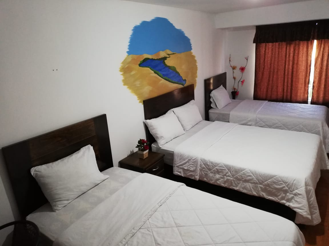 A bright room is presented with three spacious beds, each dressed in white linens. A painted mural of a desert landscape adorns the wall, adding a cultural touch. Natural light filters in through partially closed curtains, enhancing the room's welcoming atmosphere.