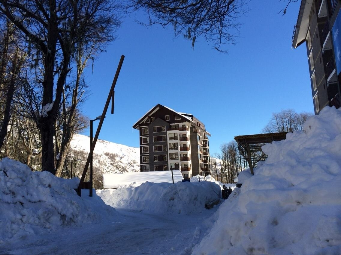 A multi-story building is set against a clear blue sky, surrounded by piles of snow. Crisp white snow blankets the ground and outlines the structure, while bare trees are visible around the area, indicating a winter landscape.