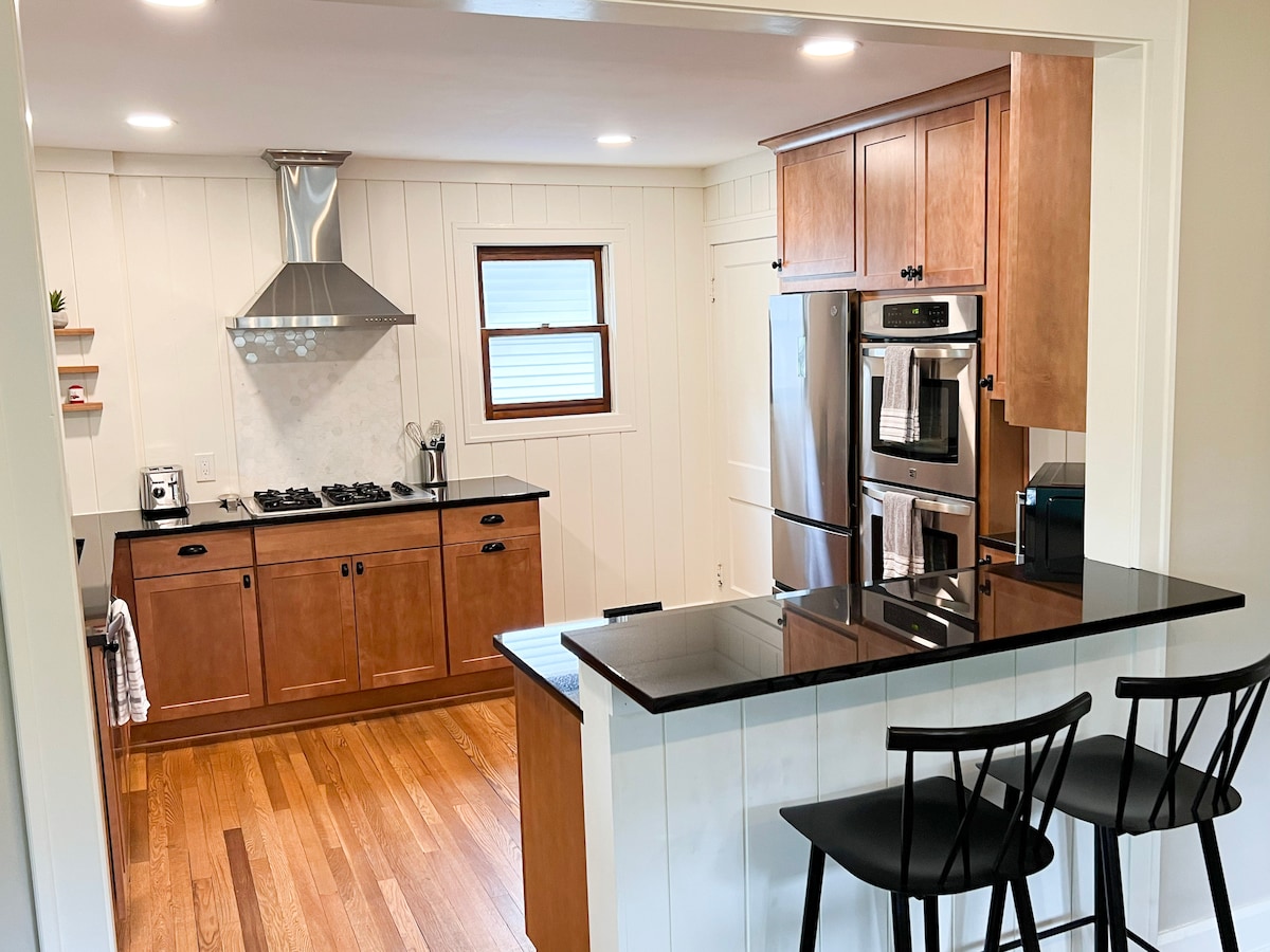 The kitchen features warm wooden cabinetry and a black countertop with bar seating for two. An overhead range hood is positioned above a gas stove, with modern appliances visible, including a refrigerator and microwave. Natural light fills the space, highlighting the elegant design.