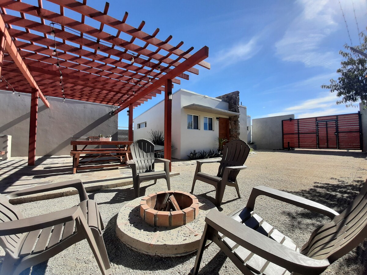An outdoor area is showcased with a pergola overhead, providing shade. A firepit surrounded by four seating chairs creates a cozy gathering space. A picnic table is visible in the background, and the property’s entrance is seen beyond the open space.