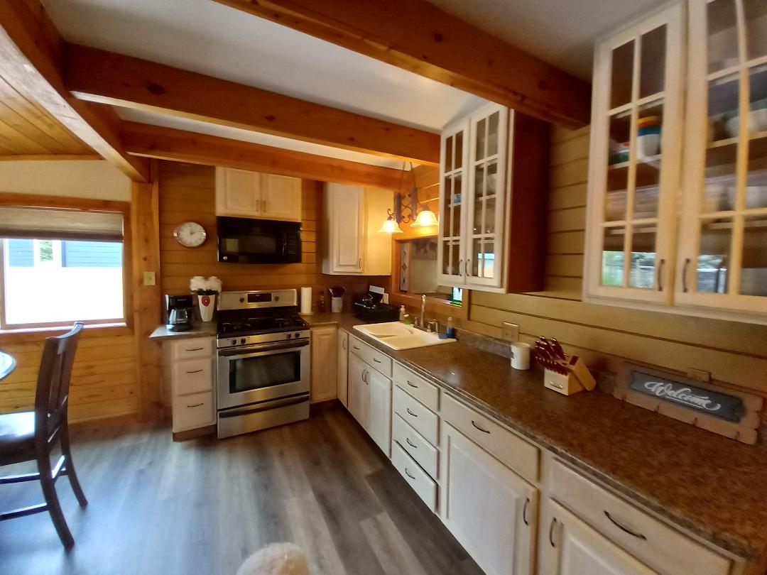 A well-equipped kitchen features wooden beams overhead and a mix of light cabinetry. Stainless steel appliances include a stove and refrigerator, and a sink is positioned beneath a window. A small dining table is visible, enhancing the functional space.