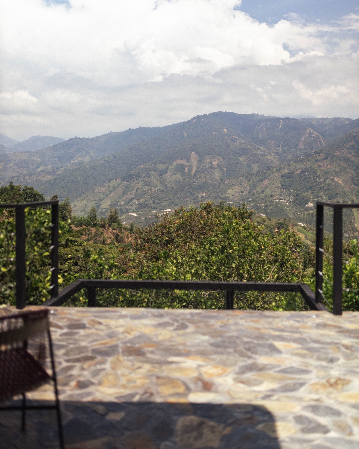 A stone patio extends outward, offering views of rolling hills covered with lush greenery under a partly cloudy sky. The surrounding landscape features varying shades of green and occasional mountain peaks in the distance.