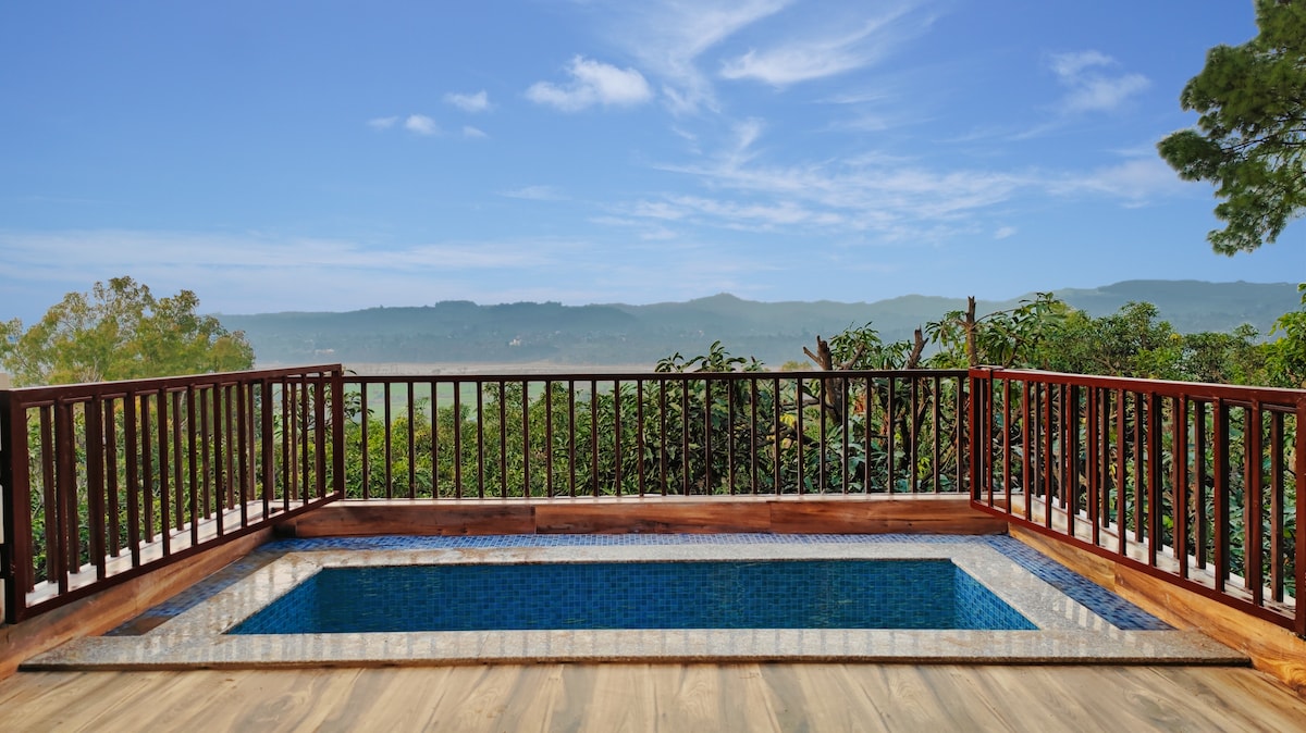 A private deck features a serene plunge pool surrounded by wooden railing. Lush greenery and distant mountains are visible beyond the space, creating a tranquil outdoor setting. The wood decking offers a warm contrast to the cool blue tiles of the pool, inviting relaxation.