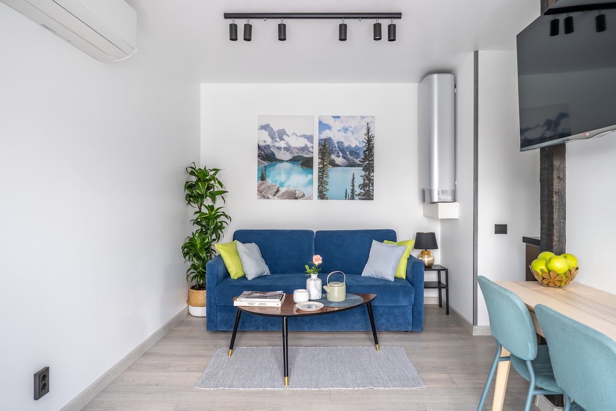 A modern living space features a blue sofa positioned in front of two landscape prints, depicting serene mountains and water. A small coffee table is adorned with a flower vase and reading materials. A dining table with light blue chairs is visible in the background.