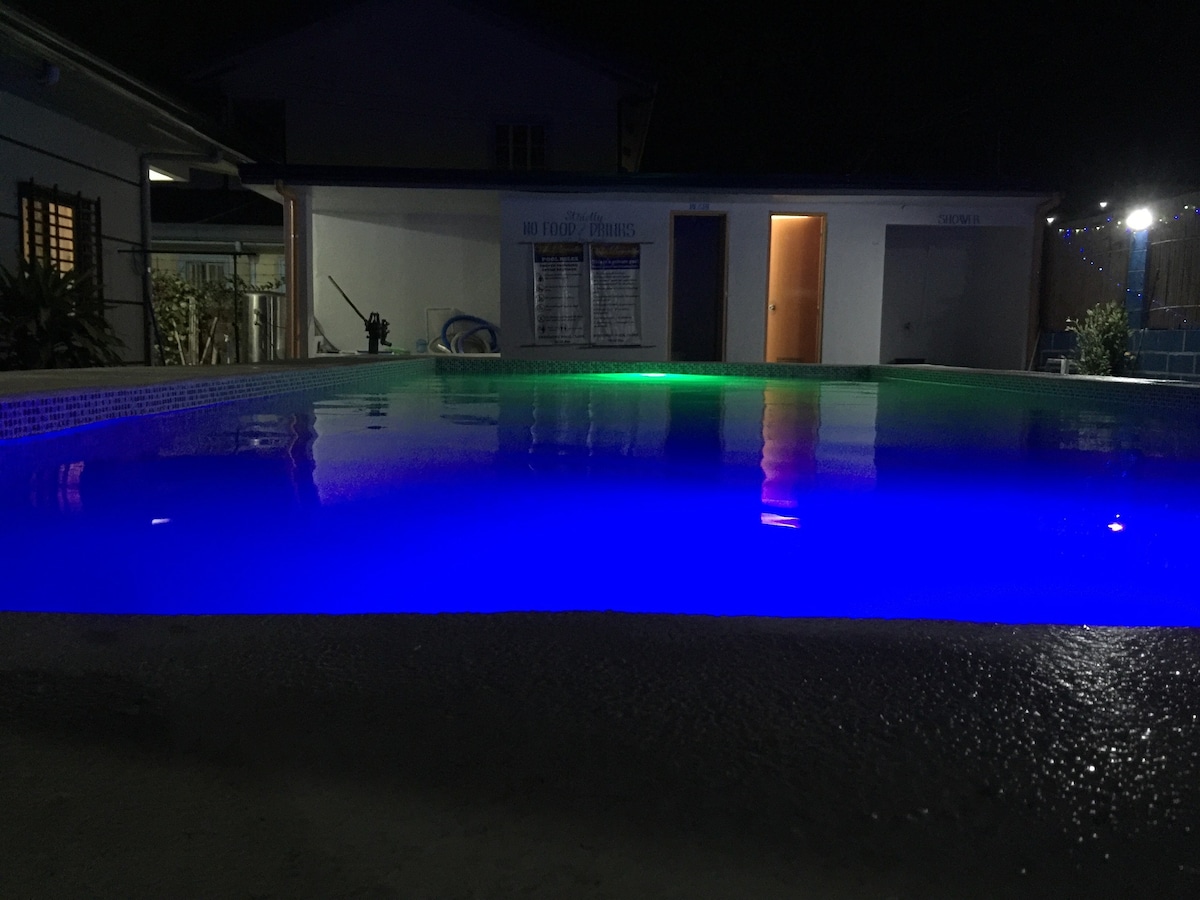 The swimming pool is illuminated at night, displaying vibrant blue and green lights that cast a calming glow over the water's surface. The surrounding area features a paved deck and is bordered by the villa's exterior, providing a private atmosphere.