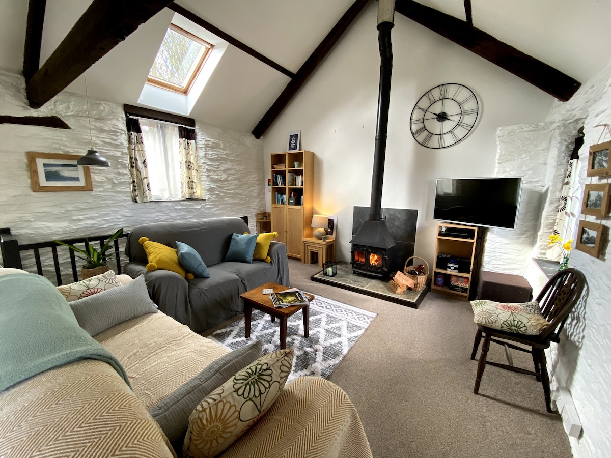 An open-plan lounge area is designed with exposed wooden beams and a high ceiling, featuring a wood-burner positioned on a black hearth. A cozy sofa and armchair are arranged around a coffee table, while a TV is mounted on a shelving unit, complemented by decorative photos on the wall.