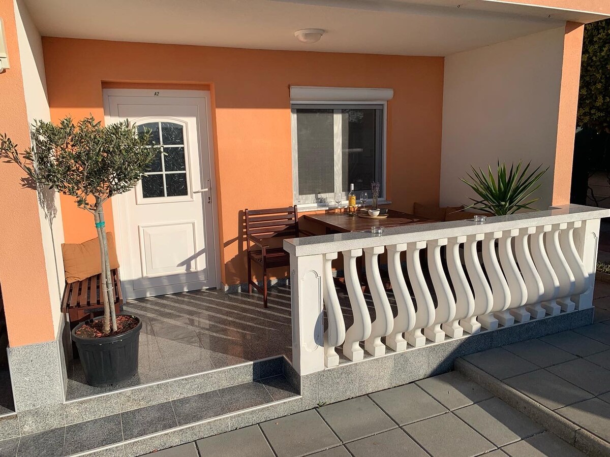 A covered terrace is featured, with a wooden table and seating arrangements for outdoor dining. Potted plants add a touch of greenery, while the entrance door leads into the apartment. The light-colored walls and ample sunlight create a welcoming ambiance.
