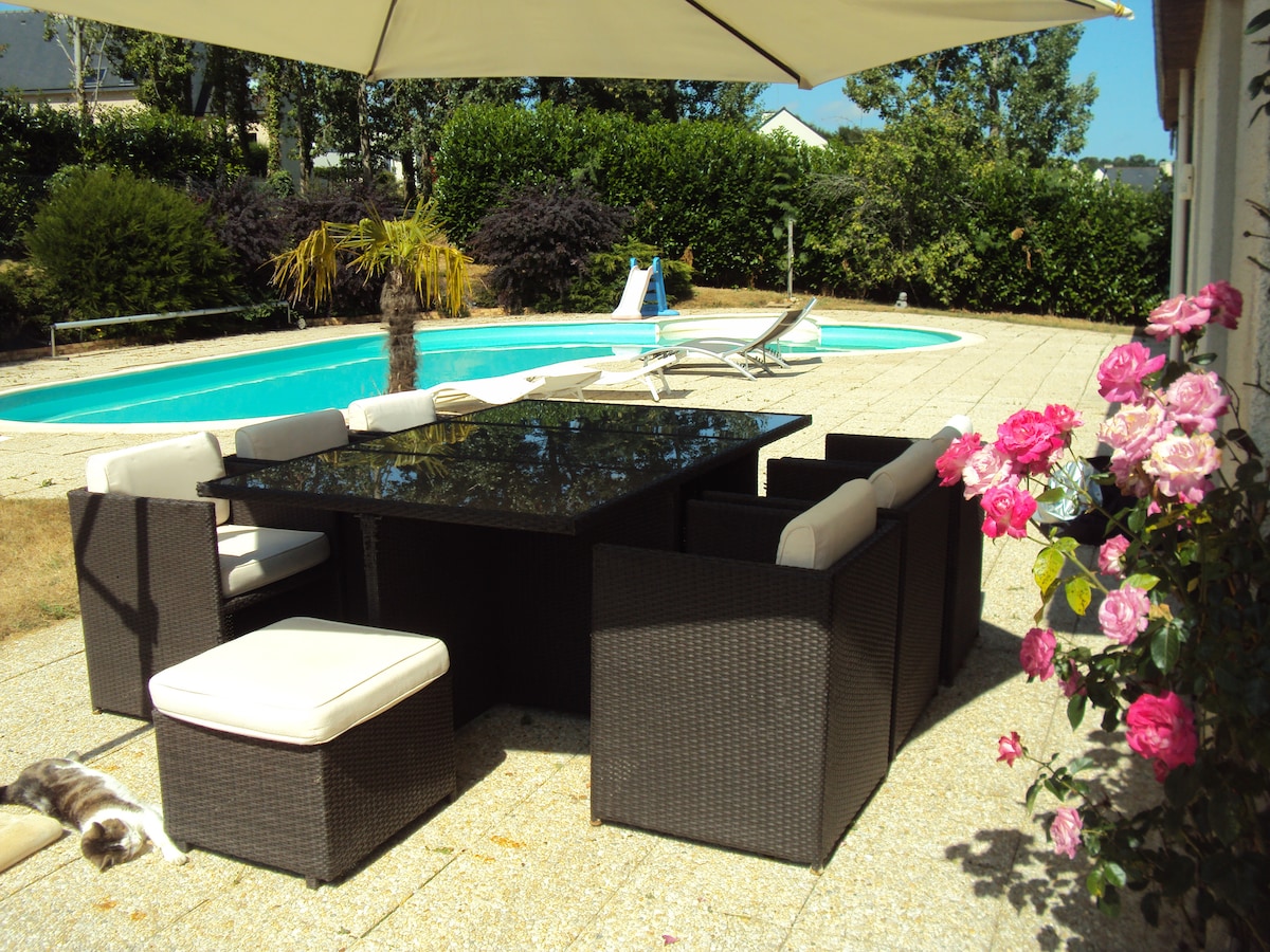 An outdoor dining area features a large table surrounded by cushioned chairs, set under a shaded umbrella. A pool lies in the background, with lounge chairs positioned beside it. Lush greenery and blooming flowers enhance the private garden setting.