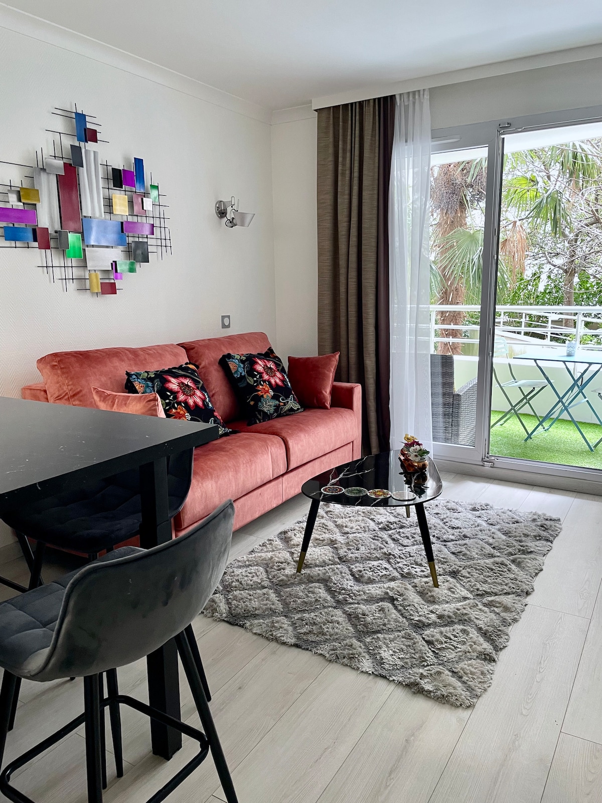 A modern living space features a coral sofa adorned with decorative pillows, paired with a sleek table and chairs. Light filters through the glass sliding doors, which open onto a small balcony. A textured rug adds warmth to the floor, enhancing the room's inviting atmosphere.