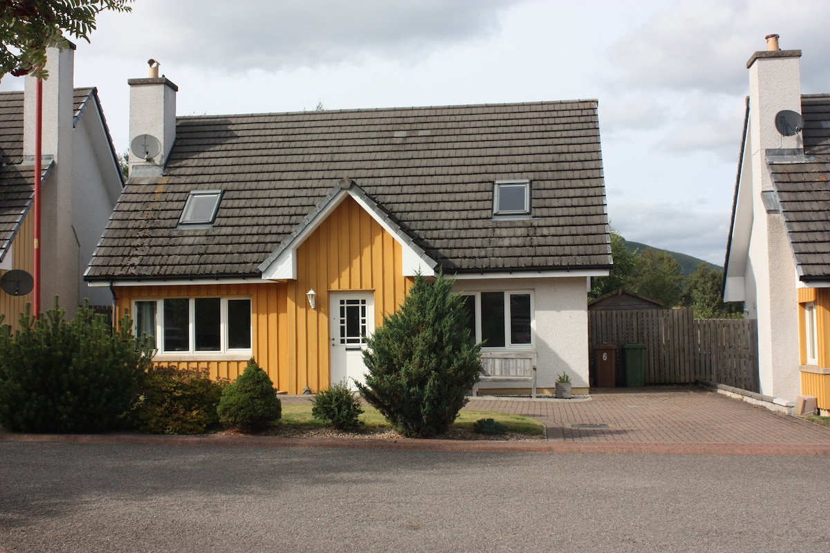The exterior of a two-story house is presented, featuring a mix of white and warm orange wood cladding. The front yard includes a neatly maintained garden with shrubs and small trees. A paved driveway leads to the entrance, with mountains visible in the background.